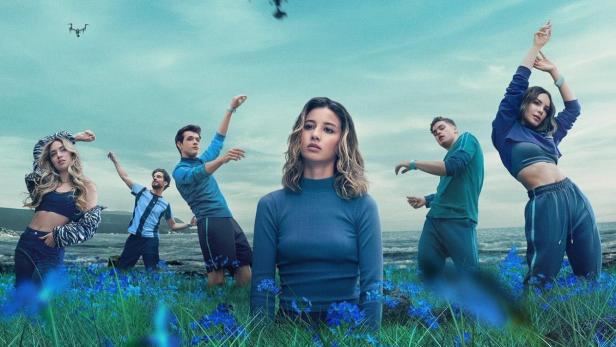 Eine Gruppe junger Leute steht auf einer Wiese unter Drohnen am Himmel.