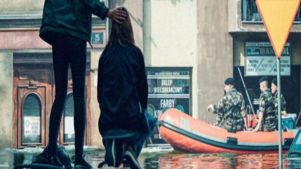 Zwei Frauen stehen auf einem Auto inmitten einer Überschwemmung; im Hintergrund ein Rettungsboot.