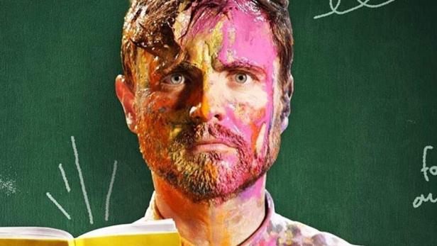 Ein Mann, mit Farbe beschmiert, hält ein Handbuch für Lehrer an Brennpunktschulen vor einer Tafel mit der Aufschrift „Almania“.