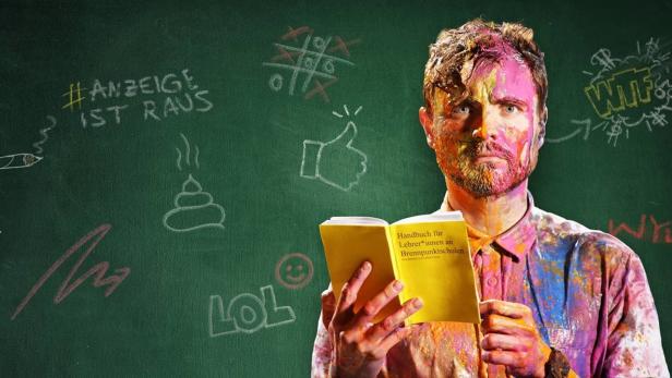 Ein mit Farbe beschmierter Mann liest ein „Handbuch für Lehrer*innen an Brennpunktschulen“ vor einer Tafel mit Kritzeleien.