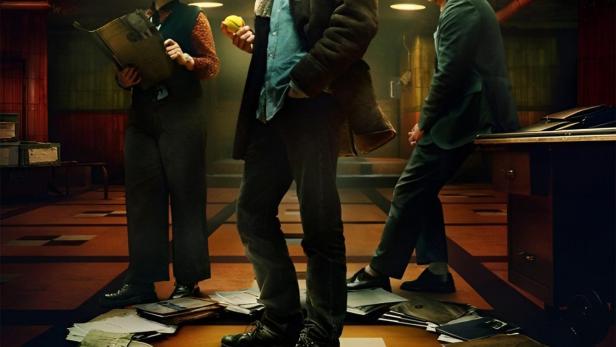 Das Filmplakat für „Dept. Q“ zeigt drei Ermittler, die in einem Büro stehen, umgeben von Akten.