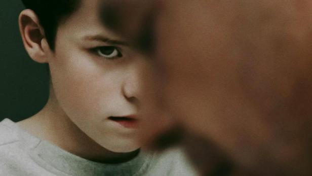 Ein ernster Junge steht vor einer verschwommenen Person; Filmtitel „Adolescence“ im Vordergrund.