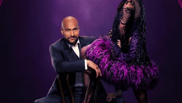 Ein Mann im Anzug sitzt auf einem Stuhl, während eine Frau im Federkleid neben ihm steht; Werbung für „Schmigadoon!“.