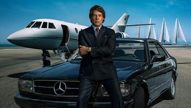 Ein Mann im Anzug posiert vor einem Privatjet, einem Oldtimer-Mercedes und Segelbooten für die Serie „Tapie“.