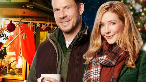 Ein Mann und eine Frau halten Tassen vor einem Weihnachtsmarkt im Film „Welcome to Christmas“.