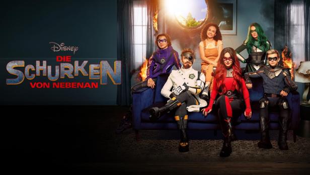 Die Besetzung der Disney+ Serie „Die Schurken von Nebenan“ posiert auf einem Sofa vor einem zerstörten Haus.