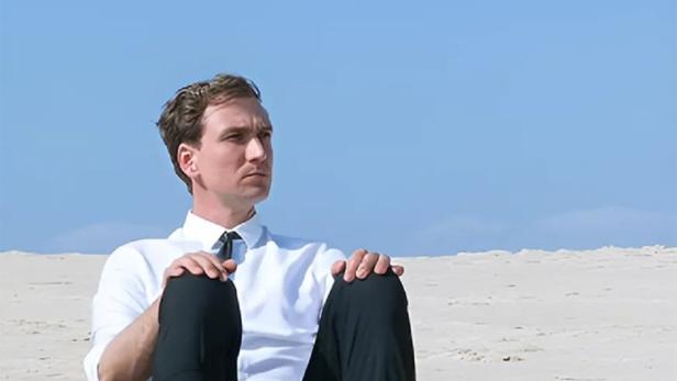 Ein Mann im Anzug sitzt barfuß am Strand, im Hintergrund das Filmplakat „Lost in Fuseta“.