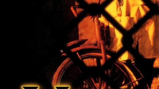 Ein Kind mit Kapuze und gelbem Regenmantel steht mit einem Fahrrad hinter einem Zaun.