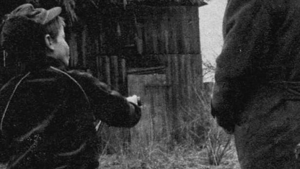 Ein Junge mit Fahrrad spricht mit einem älteren Mann vor einem Haus mit Strohdach.