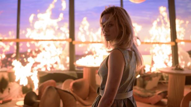 Eine junge Frau blickt über ihre Schulter, während hinter ihr ein Feuer wütet.