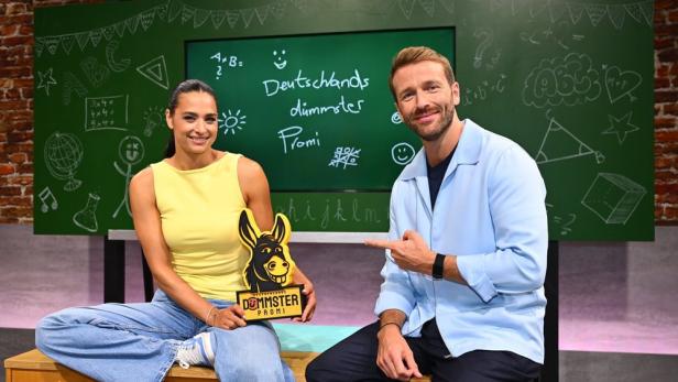 Amira Aly und Christian Düren sitzen vor einer Tafel für die Sendung „Deutschlands dümmster Promi“.