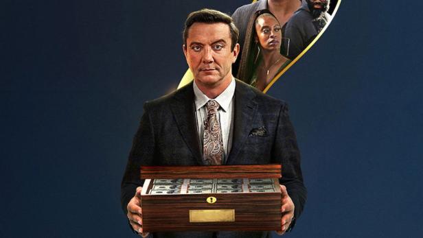 Ein Mann im Anzug hält eine Holzkiste mit Geldbündeln vor dem Hintergrund des Posters „Million Dollar Secret“.