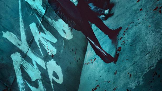 Ein Mann im Anzug steht in einem blutbefleckten Raum mit Graffiti an der Wand.