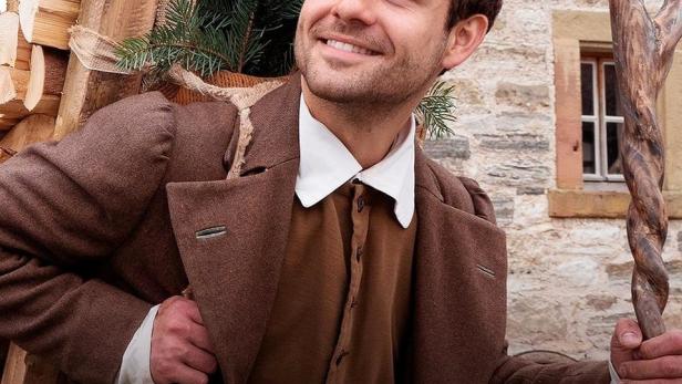 Ein lächelnder Mann in historischer Kleidung trägt Holz und Tannenzweige.