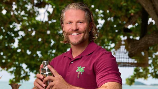 Ein lächelnder Mann, vermutlich ein Barkeeper, schüttelt einen Cocktail vor tropischer Kulisse.