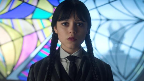Jenna Ortega als Wednesday Addams vor einem bunten Glasfenster.