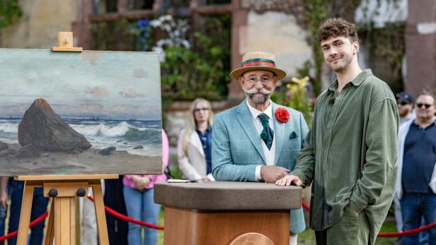 Zwei Männer stehen neben einem Gemälde mit einer Küstenlandschaft bei „Bares für Rares“.