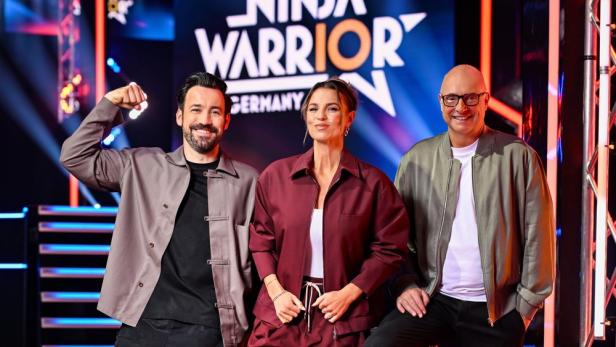 Die Moderatoren von „Ninja Warrior Germany“ posieren vor dem Logo der Show.
