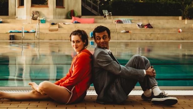 Ein junges Paar sitzt Rücken an Rücken am Beckenrand eines Freibads.