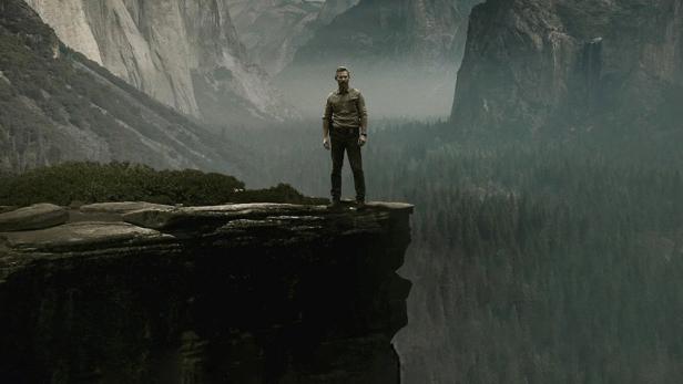 Ein Mann steht auf einem Felsvorsprung mit Blick auf eine dramatische Berglandschaft.