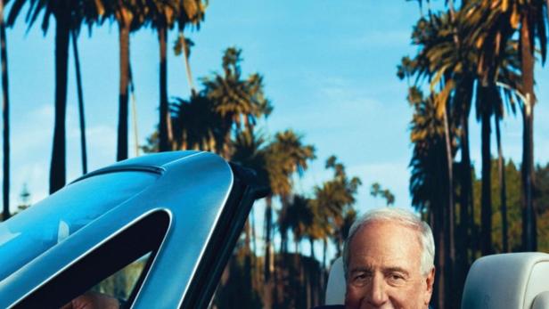 Ein Mann fährt in einem blauen Cabriolet auf einer Straße mit Palmen im Hintergrund.