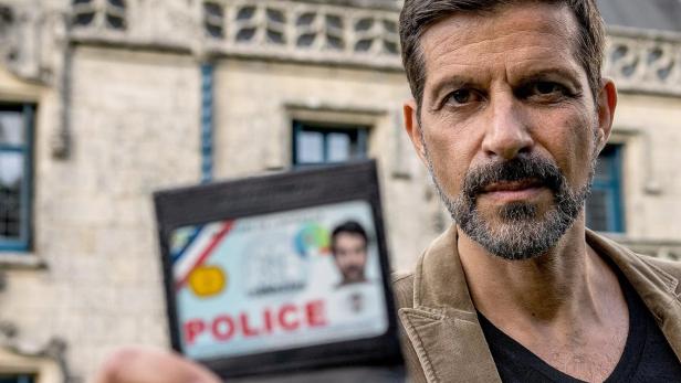 Kommissar Dupin zeigt seinen Polizeiausweis vor einem Schloss in der Bretagne.