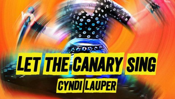 Cyndi Lauper singt in einem Konzert mit einem orangefarbenen Hintergrund.