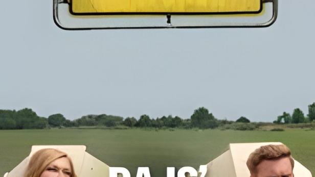 Ein Ortsschild von Osterbrarup mit dem Schriftzug „BAD“ darüber, davor zwei Personen in Holzkisten.