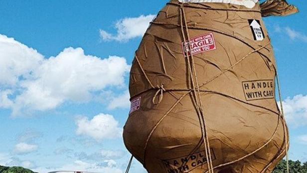 Ein übergroßer, verpackter Gartenzwerg wird auf einem Anhänger von einem Pickup-Truck transportiert.