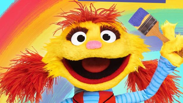 Eine gelbe, zottelige Puppe hält einen Pinsel und ein Buch mit einem Regenbogen darauf.