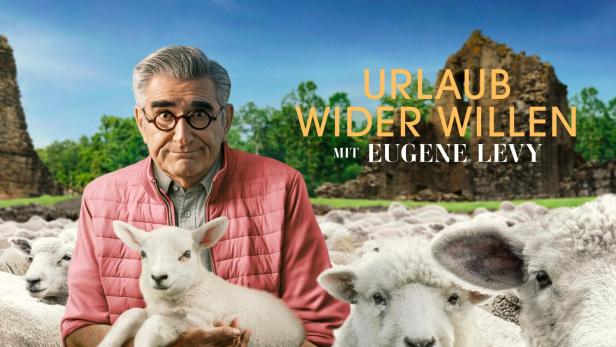 Eugene Levy hält ein Lamm vor dem Hintergrund von Schafen und einer alten Ruine für „Urlaub wider Willen“.