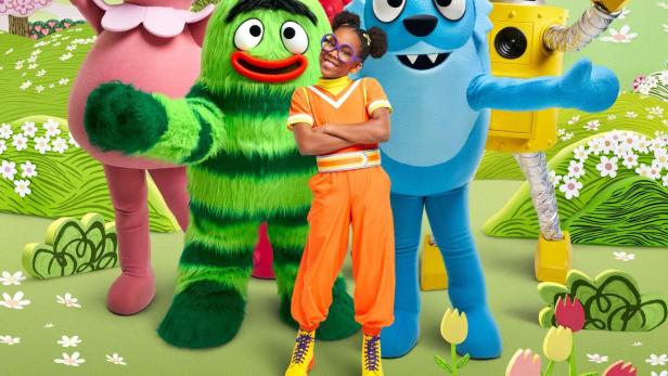 Eine junge Frau steht mit den Yo Gabba Gabba!-Charakteren vor einer grünen Landschaft.