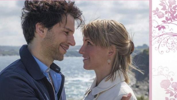 Ein lächelndes Paar steht vor dem Meer, Szene aus „Geerbtes Glück“ von Rosamunde Pilcher.