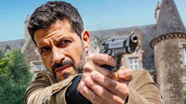 Ein Mann, Kommissar Dupin, zielt mit einer Pistole vor einem Schloss im Film „Bretonische Geheimnisse“.