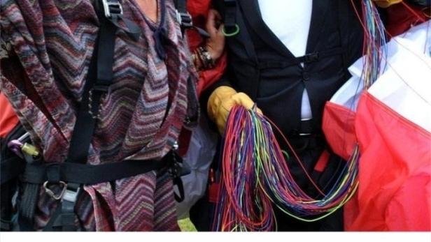 Zwei Personen mit Helmen und Fallschirmen vor einer Bergkulisse.