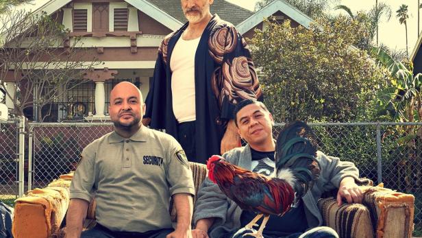 Drei Männer sitzen auf einer Couch im Freien, einer mit einem Hahn, vor dem Schriftzug „This Fool“.