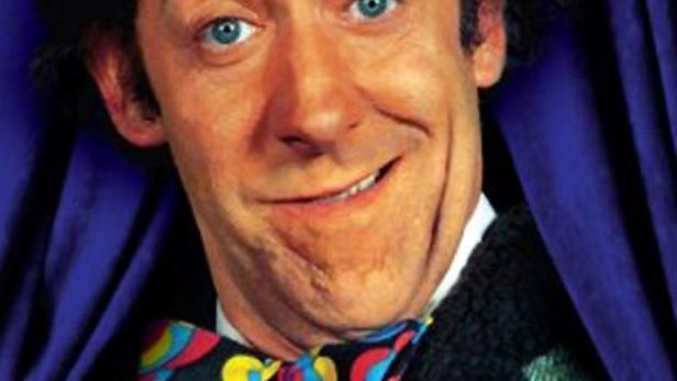 Ein Mann mit Hut und Fliege vor einem blauen Vorhang, mit dem Schriftzug „Nonstop Nonsens“.