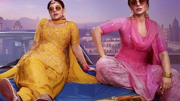 Zwei Frauen in traditioneller Kleidung sitzen auf der Motorhaube eines Autos vor einer Stadtkulisse.