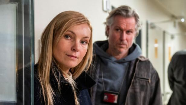 Zwei Ermittler, eine Frau mit blonden Haaren und ein Mann mit grauem Haar, stehen in einem Flur.