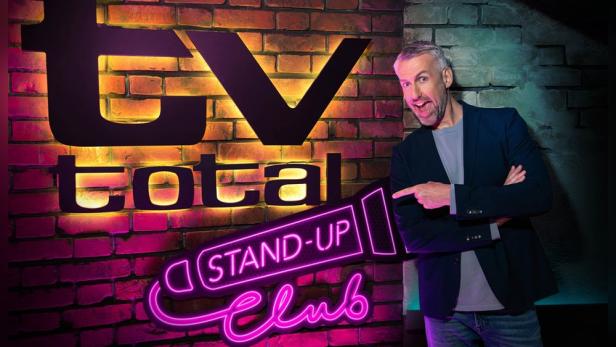 Ein Mann steht vor dem „TV Total Stand-Up Club“-Logo auf einer Backsteinmauer.