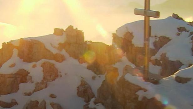 Ein Gipfelkreuz auf einem schneebedeckten Berg in den Alpen bei Sonnenaufgang.