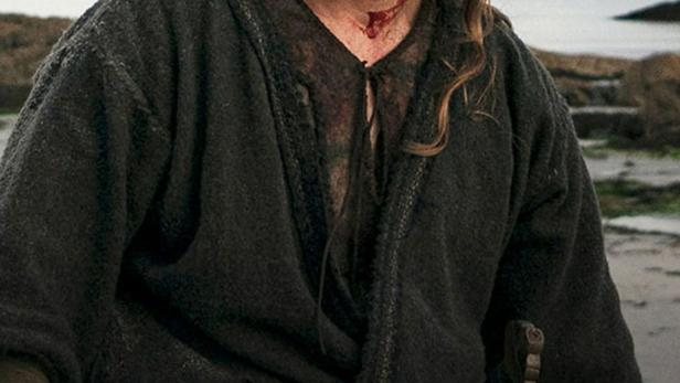 Ein blutverschmierter Wikinger steht vor einer Küstenlandschaft.