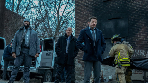 Jeremy Renner steht in seiner Serienrolle als „Mayor of Easttown“ vor einer dunklen Straßenecke, wo es offenbar gebrannt hat.