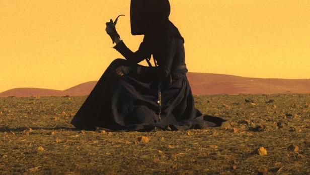 Eine in Schwarz gekleidete Person sitzt in einer Wüstenlandschaft und raucht eine Pfeife.