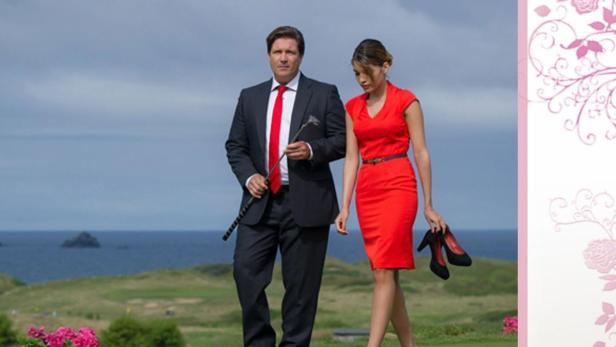 Ein Mann im Anzug und eine Frau im roten Kleid gehen barfuß auf einem Golfplatz am Meer entlang.