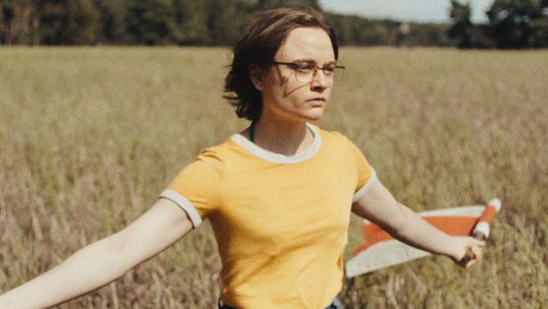 junge Frau mit Brille in gelbem Shirt steht mit ausgebreiteten Armen vor einem Kornfeld.