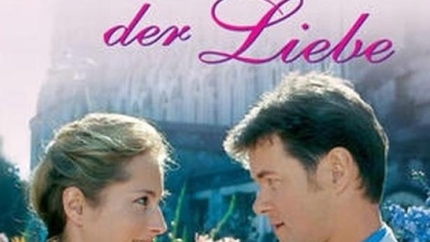 Ein Paar steht vor Blumen und dem Titelbild von Rosamunde Pilchers „Flamme der Liebe“.