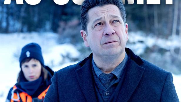 Ein Mann in einem Mantel schreibt Notizen, während eine Polizistin im Hintergrund steht.