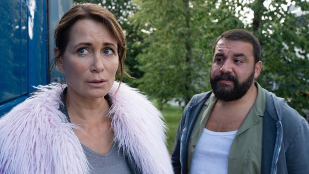 TV-Filmszene: Frau steht in Parkgebiet vor Fahrerkabine eines Trucks und rechts von ihr blickt der vollbärtige Trucker auf sie.