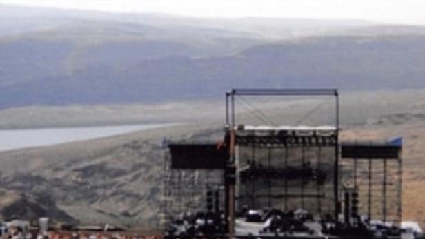 Eine große Menschenmenge versammelt sich vor einer Bühne im Freien mit Blick auf eine hügelige Landschaft.